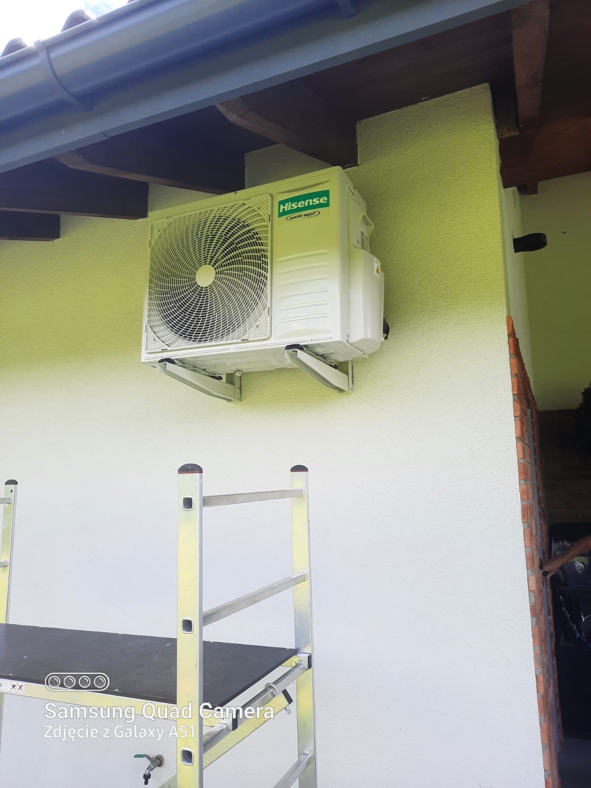 Biała jednostka zewnętrzna klimatyzacji Hisense zamontowana na wspornikach na elewacji budynku, widoczny fragment rusztowania aluminiowego.
