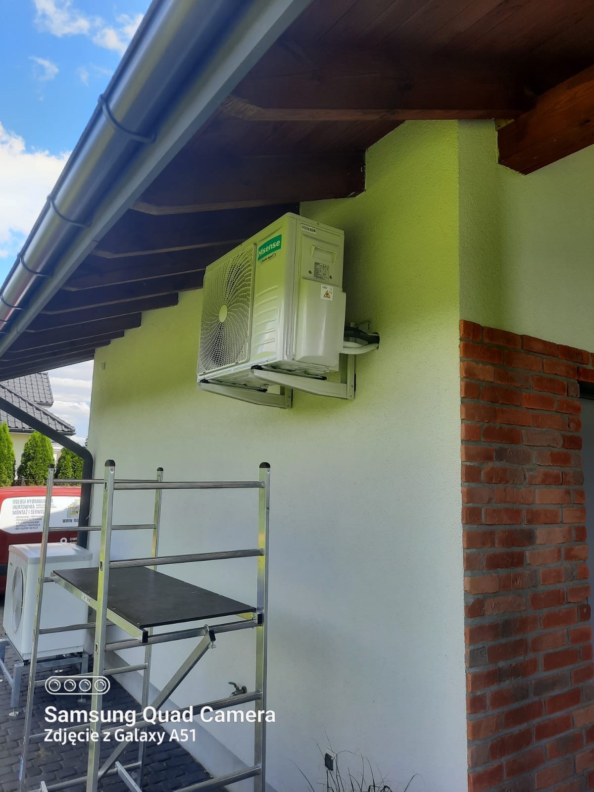 Biała jednostka zewnętrzna klimatyzacji Hisense zamontowana na jasnej elewacji budynku, widoczna drabina z platformą, czerwona cegła dekoracyjna na rogu budynku i fragment rynny.