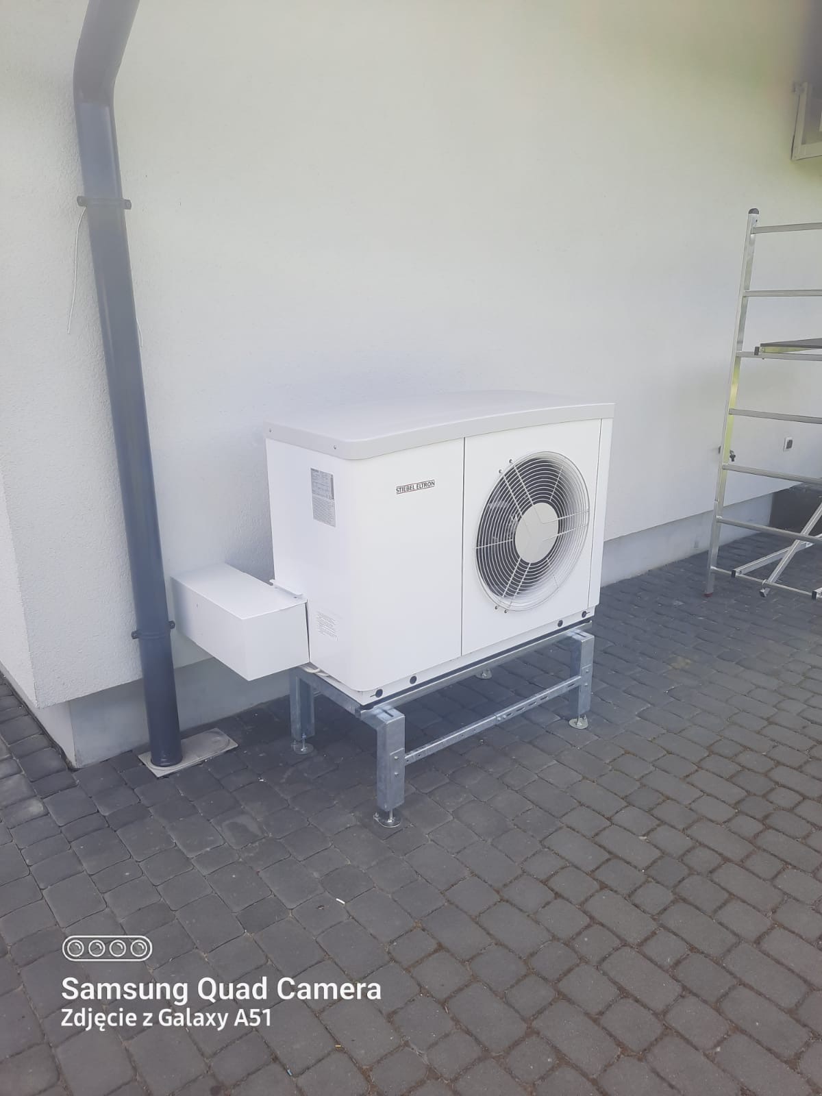 Biała pompa ciepła marki Stiebel Eltron zamontowana na metalowej konstrukcji przy białej ścianie budynku, obok rynny spustowej i drabiny.