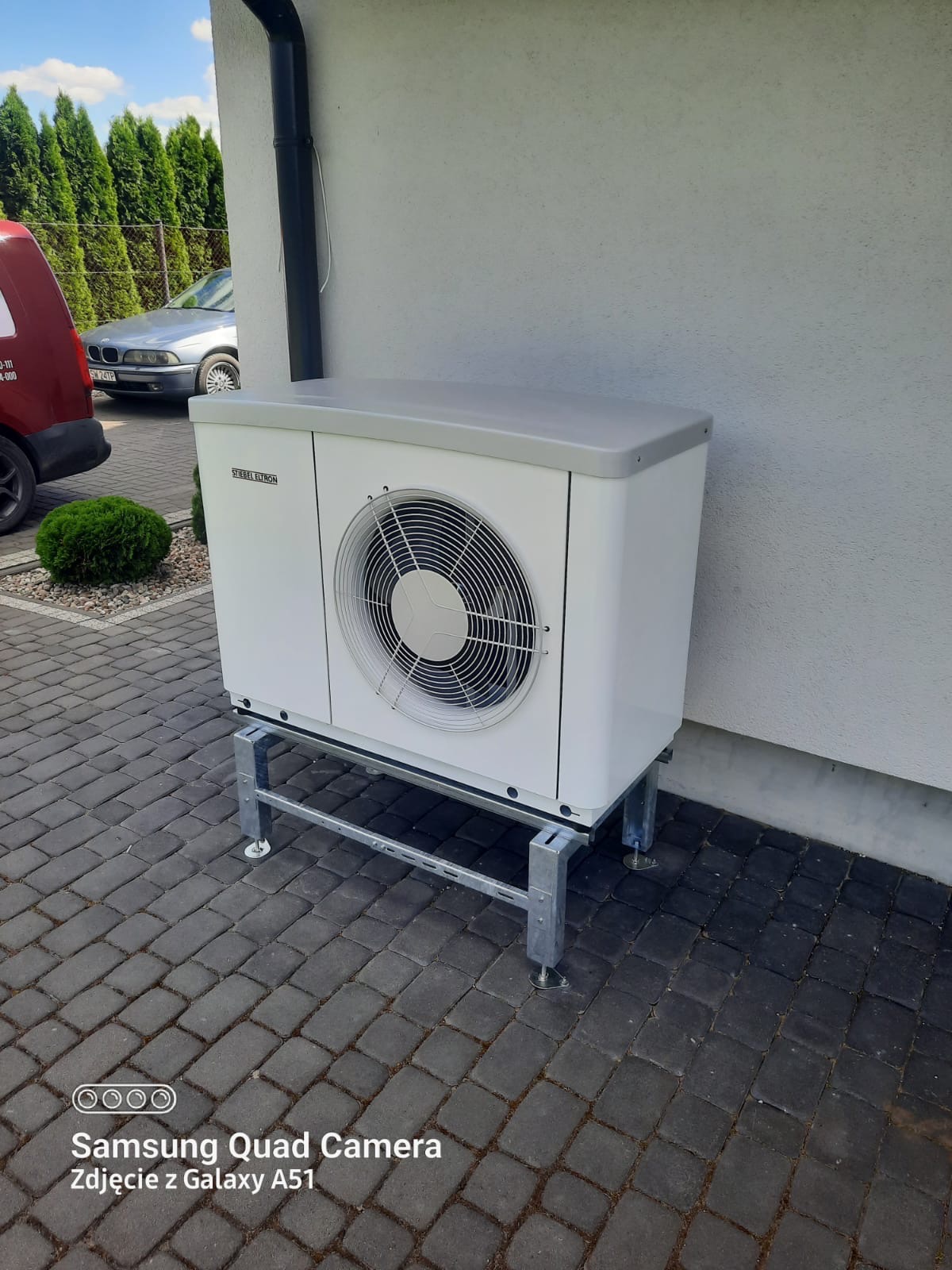 Biała pompa ciepła Stiebel Eltron zamontowana na metalowej konstrukcji na kostce brukowej, widoczny fragment elewacji budynku i zaparkowany samochód