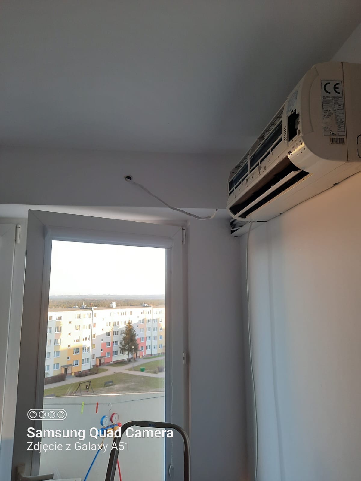 Biały klimatyzator ścienny zamontowany w rogu pokoju, widoczny kabel wychodzący ze ściany, okno z widokiem na blok mieszkalny w tle.