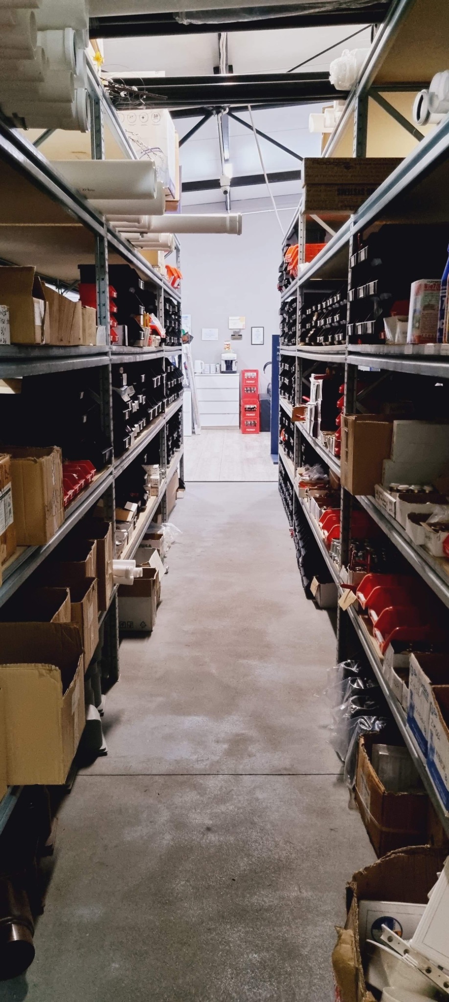 Magazyn instalacyjny z rzędami metalowych regałów wypełnionych rurami, złączkami i akcesoriami hydraulicznymi, widok w głąb jasnego pomieszczenia.