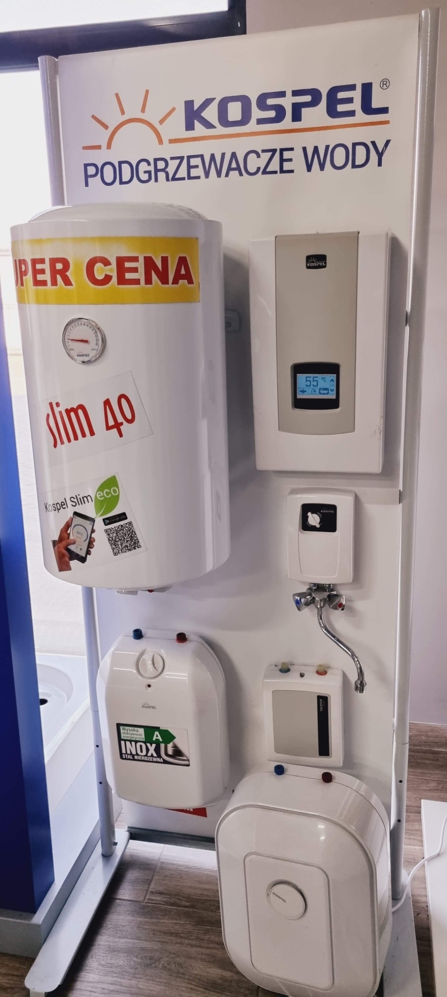 Ekspozycja różnych modeli podgrzewaczy wody marki Kospel na białym stojaku, w tym zbiornikowy podgrzewacz 'Slim 40' z naklejką 'Super Cena' i informacją o sterowaniu mobilnym, oraz przepływowe...