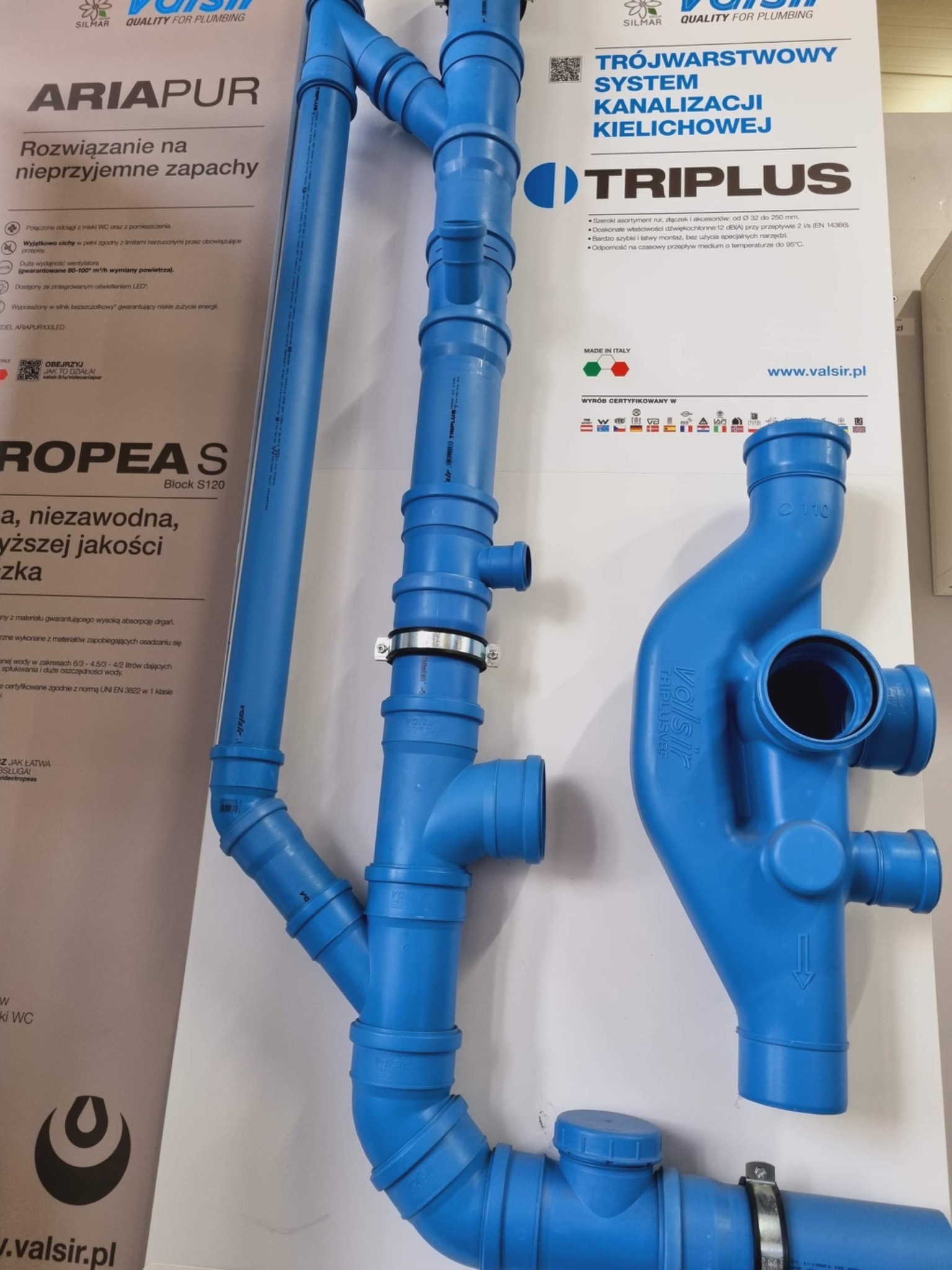 Niebieskie rury kanalizacyjne z kielichami i złączkami, prezentowane obok materiałów reklamowych firmy Valsir, widoczne napisy Ariapur i Triplus, metalowe obejmy zaciskowe.