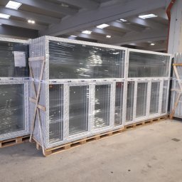 FAMM ITALIA SP.ZO.O - Magazynowane okna PCV, zabezpieczone folią i drewnianymi wspornikami, ułożone na paletach.