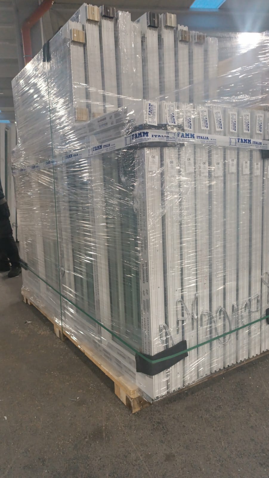 Paleta z zapakowanymi w folię białymi profilami okiennymi, zabezpieczona taśmami, widoczny fragment nogi osoby w kombinezonie roboczym.