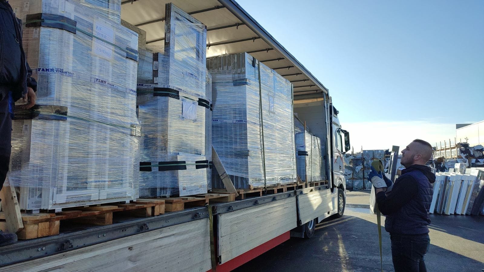 Załadunek okien na paletach, owiniętych folią stretch, na naczepę ciężarówki; pracownik zabezpiecza ładunek pasami transportowymi.