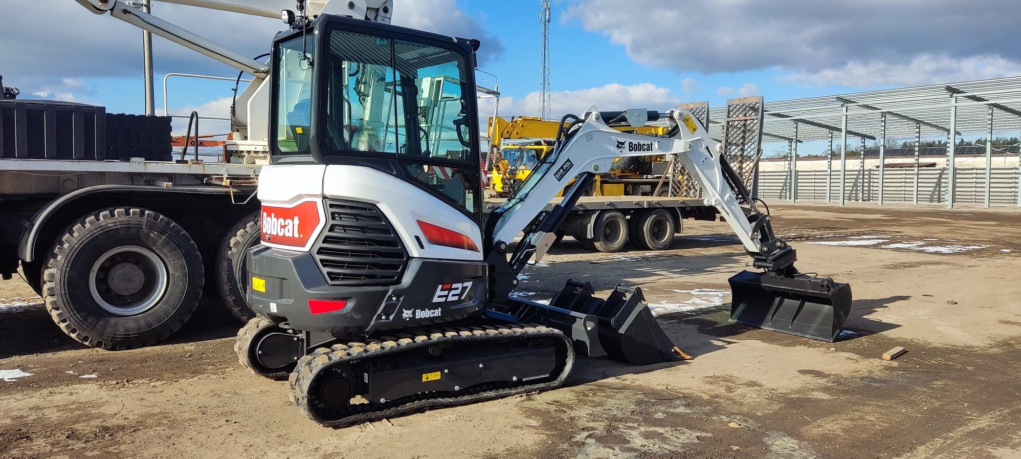 Biało-czarna minikoparka Bobcat E27 na placu budowy, widok z boku, z łyżkami skierowanymi w stronę widza. W tle inne maszyny i konstrukcja hali.