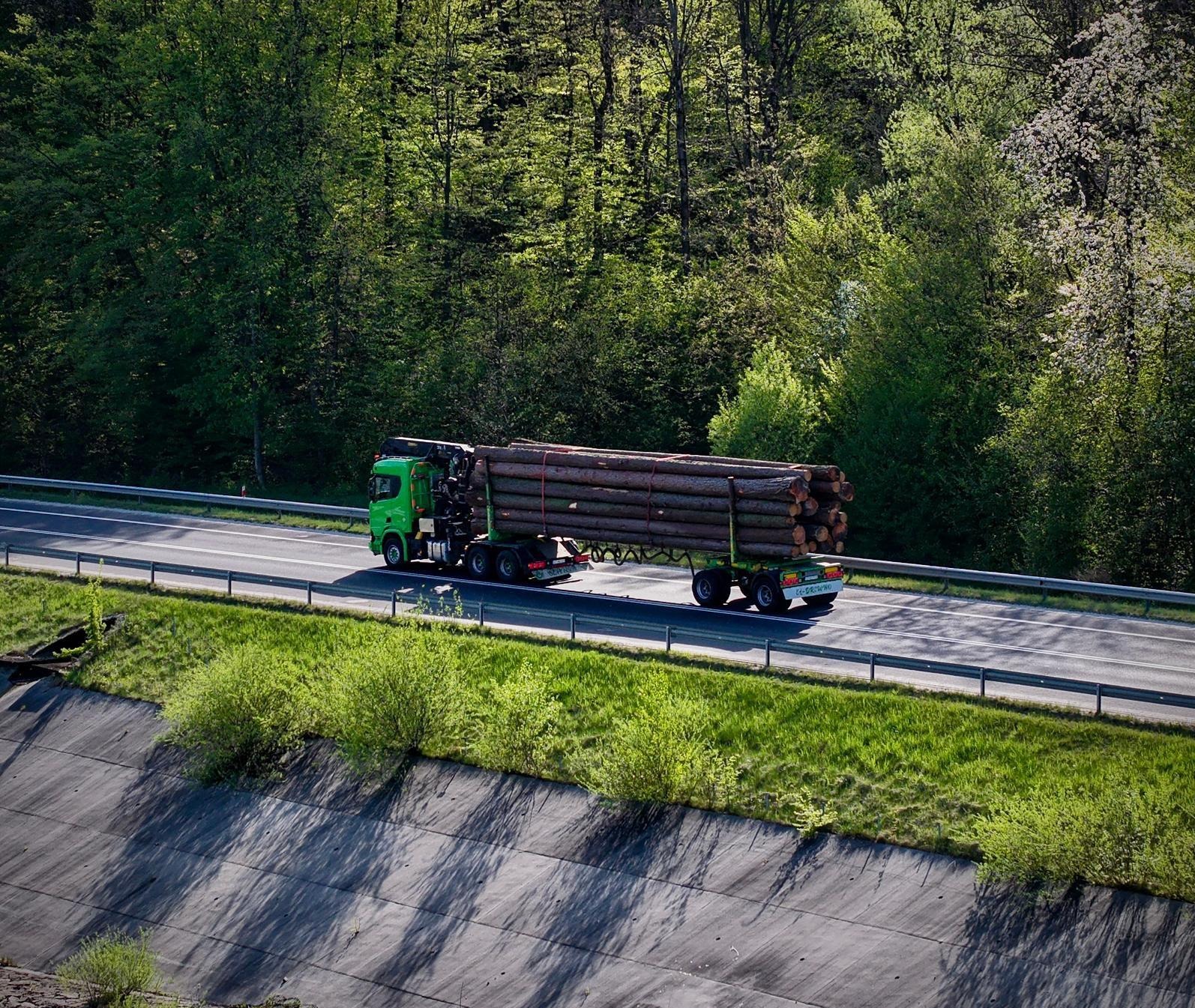 Zielona ciężarówka z drewnem na autostradzie, otoczona zielenią, w słoneczny dzień. Widok z góry na pojazd w ruchu, z widocznym ładunkiem drewna.