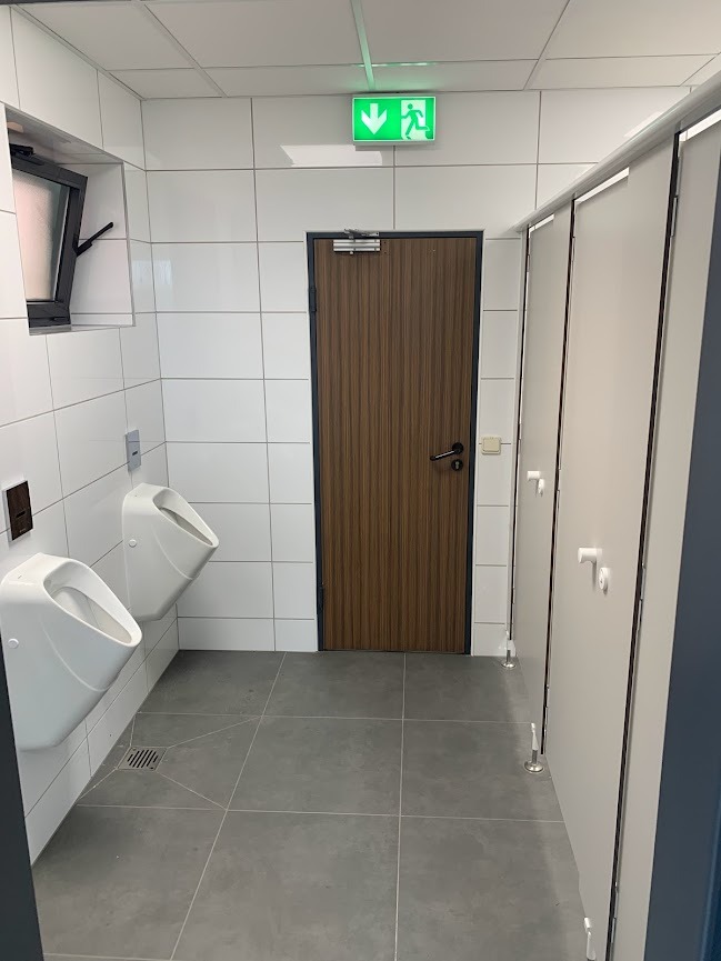 Publiczna toaleta z dwoma pisuarami, brązowymi drzwiami z napisem 'wyjście' oraz kabinami toaletowymi z białymi drzwiami i szarą podłogą z płytek.