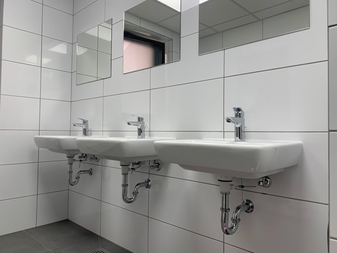 Rząd trzech identycznych, białych umywalek zamontowanych na ścianie z białych, kwadratowych płytek, każda z chromowaną baterią i syfonem, pod nimi szara podłoga z kratką ściekową.