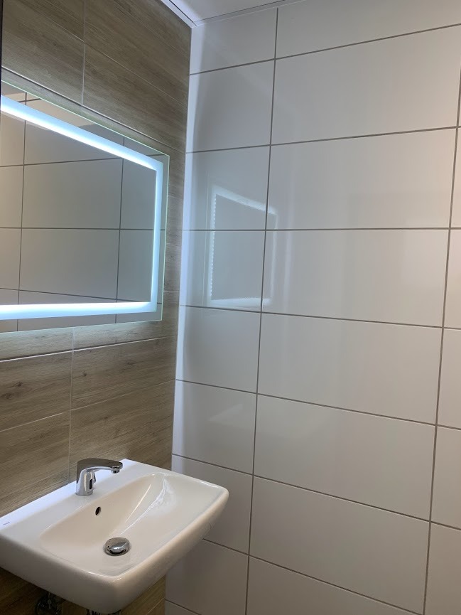 Biała umywalka z chromowaną baterią i odpływem na tle ściany wyłożonej białymi, błyszczącymi płytkami ceramicznymi i drewnopodobnymi płytkami, oświetlona lustrem z podświetleniem LED.