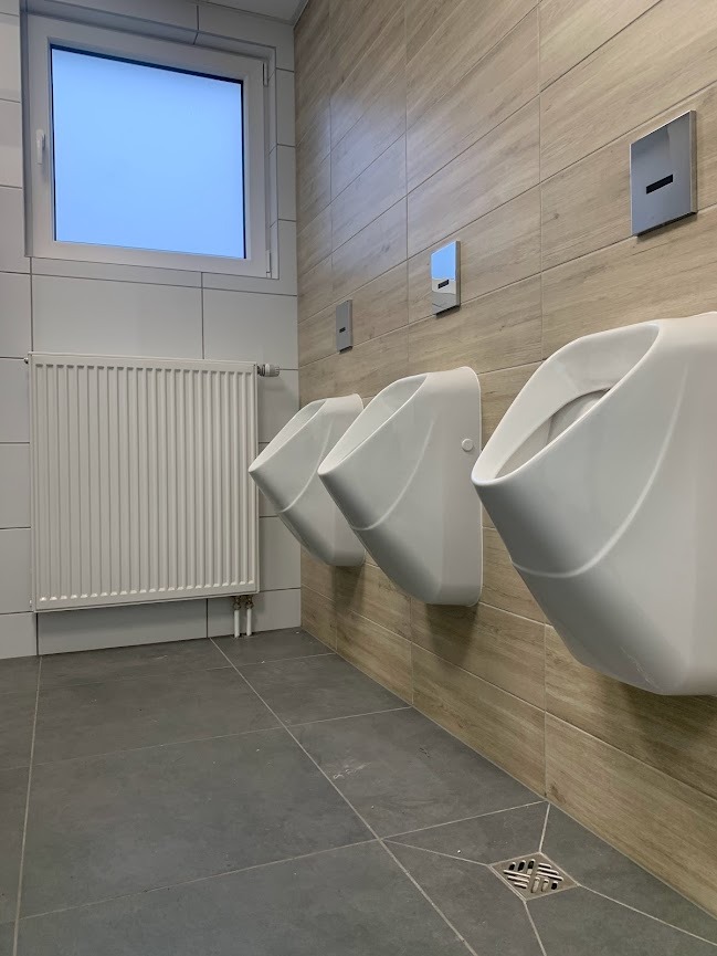 Rząd trzech białych pisuarów zamontowanych na ścianie wyłożonej płytkami imitującymi drewno w publicznej toalecie, z szarymi płytkami podłogowymi i grzejnikiem żeberkowym po lewej stronie.