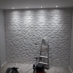 Krys-Mat - Ściana wykończona białymi panelami imitującymi cegłę, oświetlona punktowymi lampami sufitowymi. Widoczna drabina, odkurzacz i narzędzia malarskie na podłodze z paneli.