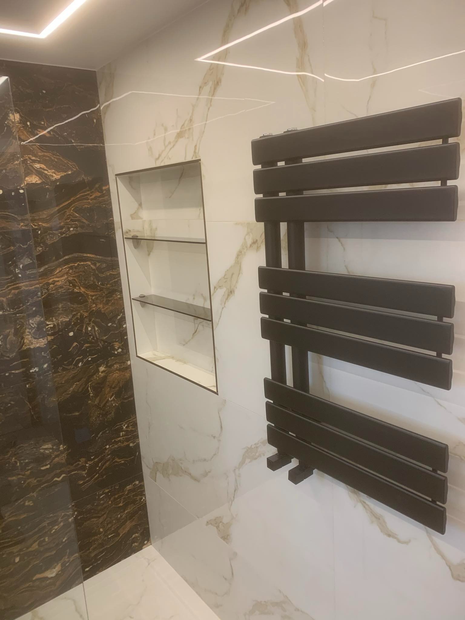 Nowoczesna łazienka z marmurowymi płytkami w odcieniach bieli i brązu, podświetlana półka wnękowa z szklanymi półkami oraz czarny, drabinkowy grzejnik.