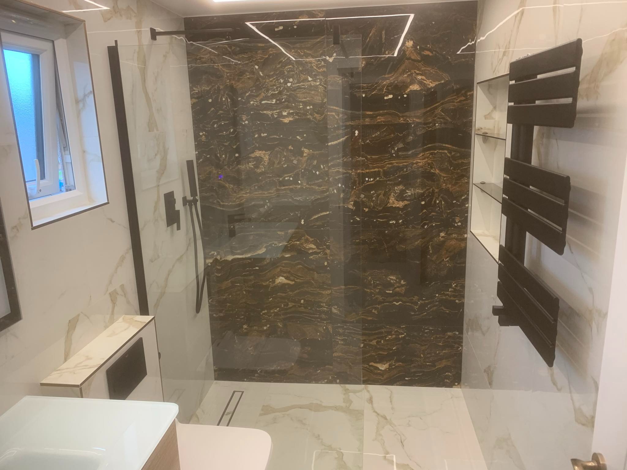 Nowoczesna łazienka z kabiną prysznicową typu walk-in, wykończona płytkami imitującymi marmur w odcieniach bieli i brązu, z czarną armaturą i grzejnikiem drabinkowym.