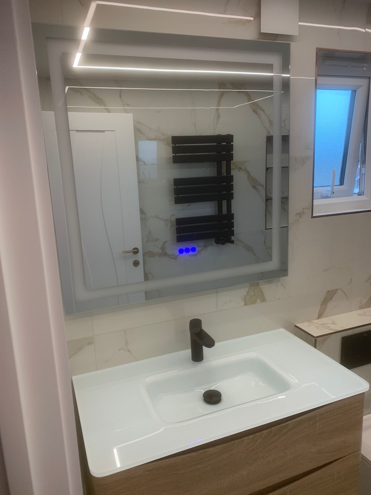 Nowoczesna łazienka z podświetlanym lustrem, umywalką z szafką z drewnopodobnym frontem i grzejnikiem drabinkowym w odbiciu lustra.