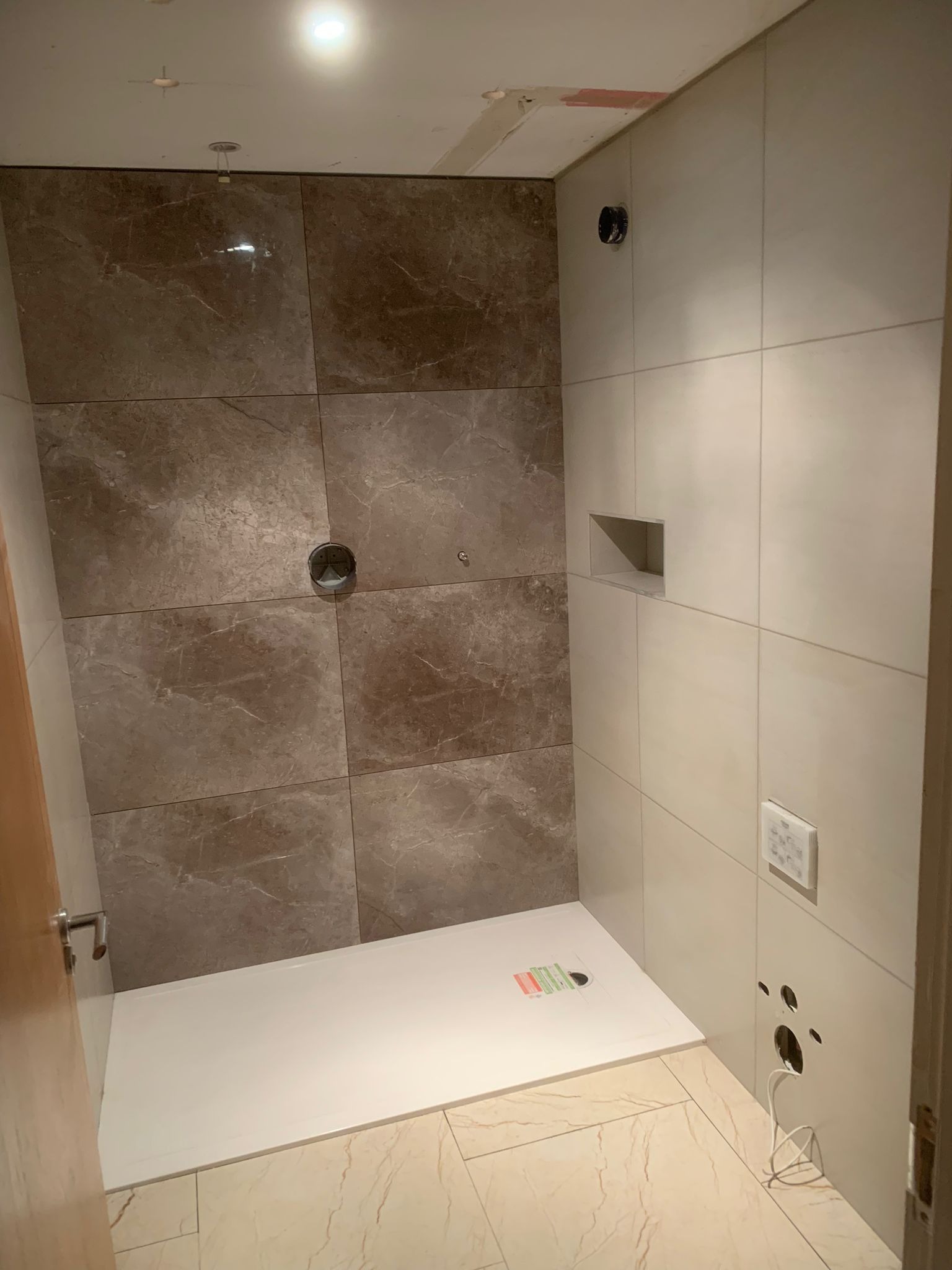 Niedokończona łazienka z brązowymi i kremowymi płytkami, biały brodzik, otwory na armaturę, brakujące wykończenia sufitu i wystające przewody.