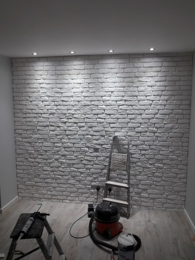 Ściana wykończona białymi panelami imitującymi cegłę, oświetlona punktowymi lampami sufitowymi. Widoczna drabina, odkurzacz i narzędzia malarskie na podłodze z paneli.