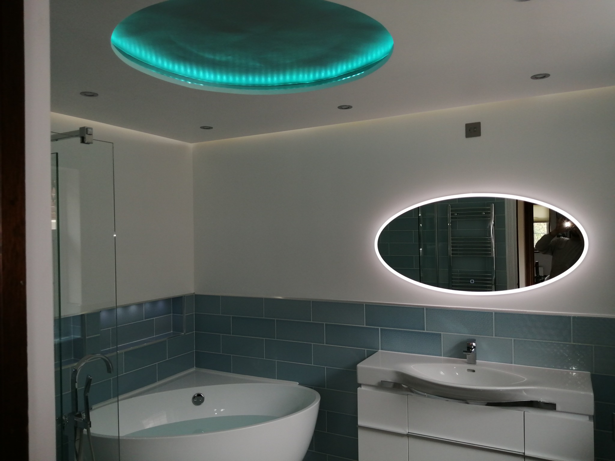 Nowoczesna łazienka z okrągłym, podświetlanym sufitem w kolorze turkusowym, owalnym lustrem z podświetleniem LED, wanną z hydromasażem i umywalką na szafce, wykończona niebieską glazurą.