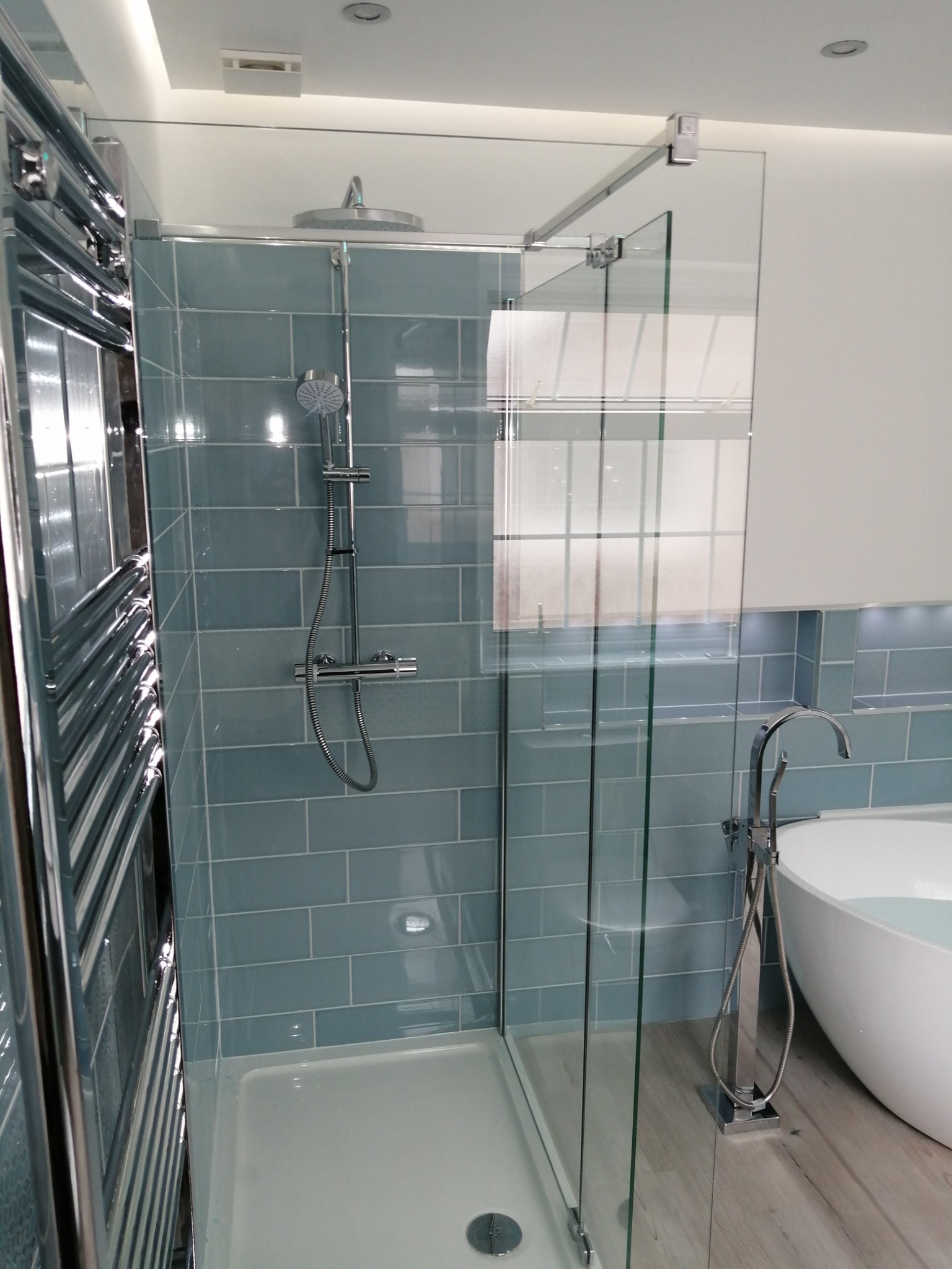 Nowoczesna łazienka z kabiną prysznicową typu walk-in, błękitnymi płytkami ułożonymi w cegiełkę, grzejnikiem drabinkowym i wolnostojącą wanną wypełnioną wodą.