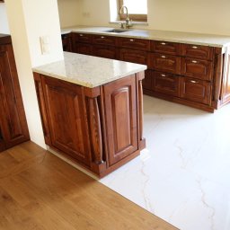 Usługi stolarskie Skupień - Klasyczna kuchnia z ciemnego drewna z blatem z jasnego granitu, widok na wyspę kuchenną i zabudowę przy ścianie, białe płytki podłogowe imitujące marmur, parkiet dębowy w jodełkę.