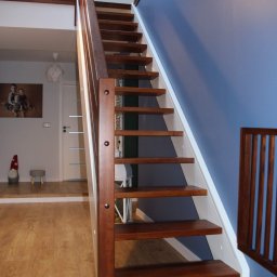 Usługi stolarskie Skupień - Nowoczesne, drewniane schody z ciemnego drewna z białymi elementami konstrukcyjnymi, widoczne z dołu, z balustradą na górze, na tle błękitnej ściany i jasnego wnętrza.