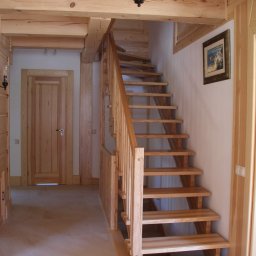 Usługi stolarskie Skupień - Jasne, drewniane schody z balustradą w domu o drewnianym wykończeniu, widoczne drzwi z drewnianą ramą, podłoga wyłożona jasnymi płytkami, na ścianie obraz w złotej ramie i lampa wisząca w stylu retro.