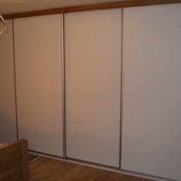 Usługi stolarskie Skupień - Czterodrzwiowa szafa przesuwna z białymi frontami i drewnianym wykończeniem, widoczna z perspektywy łóżka w sypialni, z ozdobną lampą wiszącą i zasłoną w kwiaty.