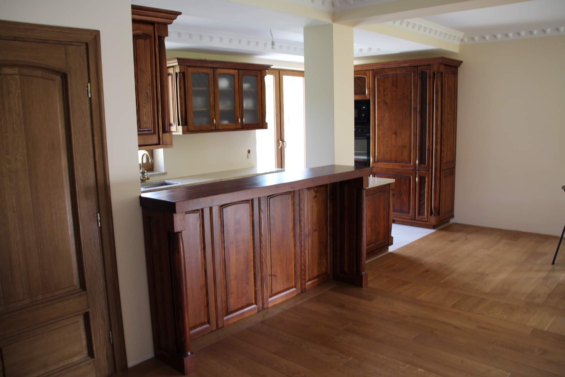 Klasyczna, drewniana kuchnia z wyspą i szafkami wiszącymi z przeszklonymi frontami, widok z perspektywy podłogi, z widocznymi detalami wykończenia sufitu.