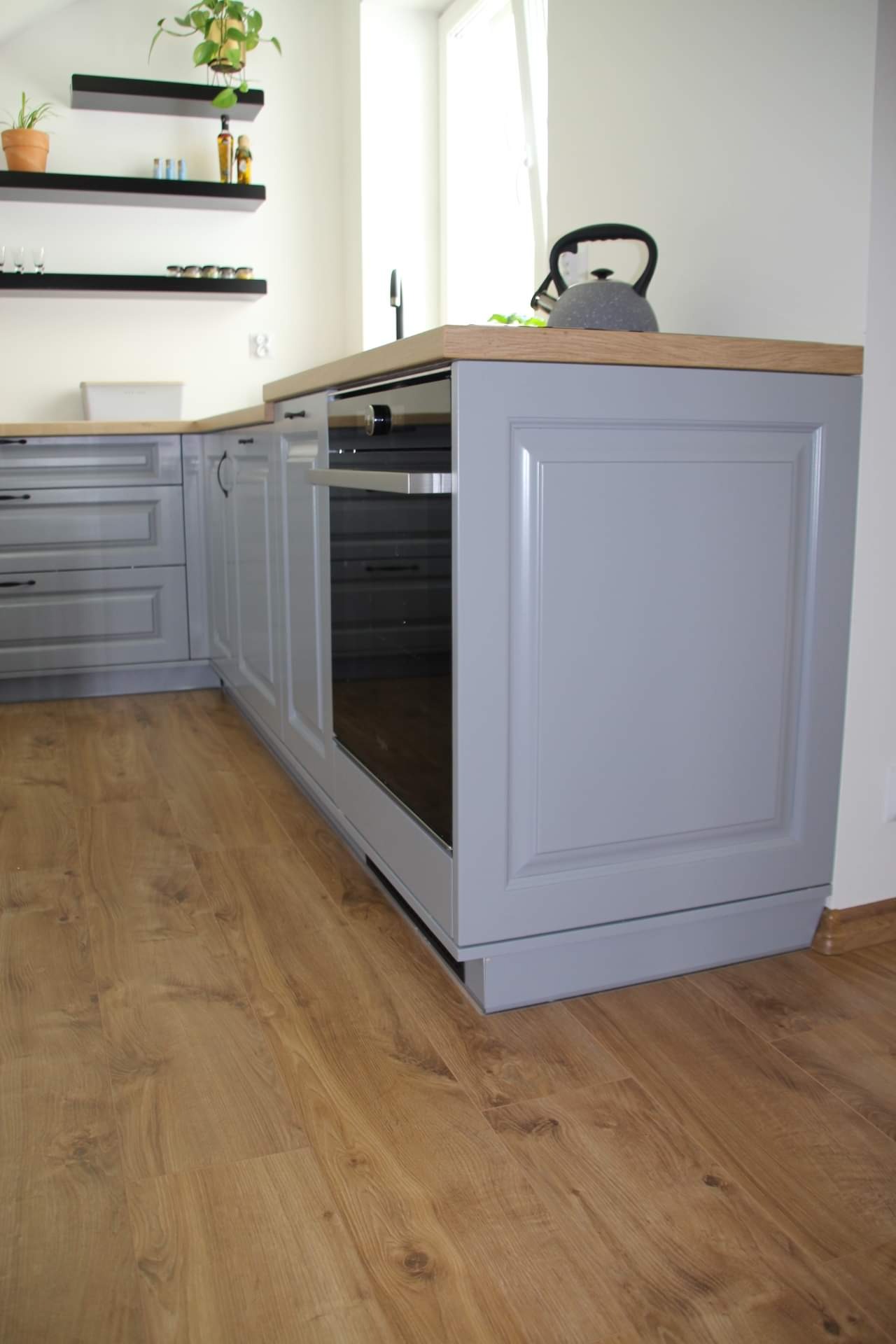 Szara zabudowa kuchenna z piekarnikiem, drewniany blat, podłoga z paneli, minimalistyczne półki na ścianie z roślinami doniczkowymi, czajnik na blacie.