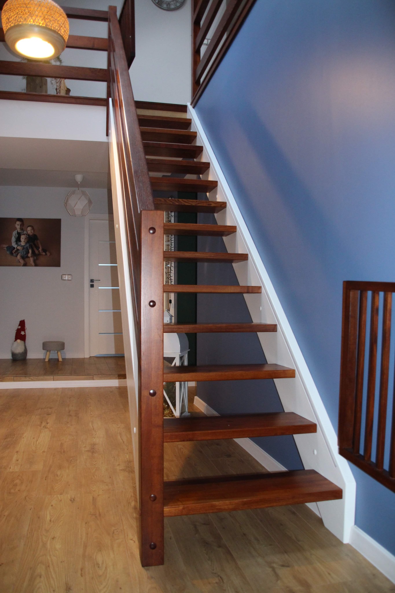 Nowoczesne, drewniane schody z ciemnego drewna z białymi elementami konstrukcyjnymi, widoczne z dołu, z balustradą na górze, na tle błękitnej ściany i jasnego wnętrza.