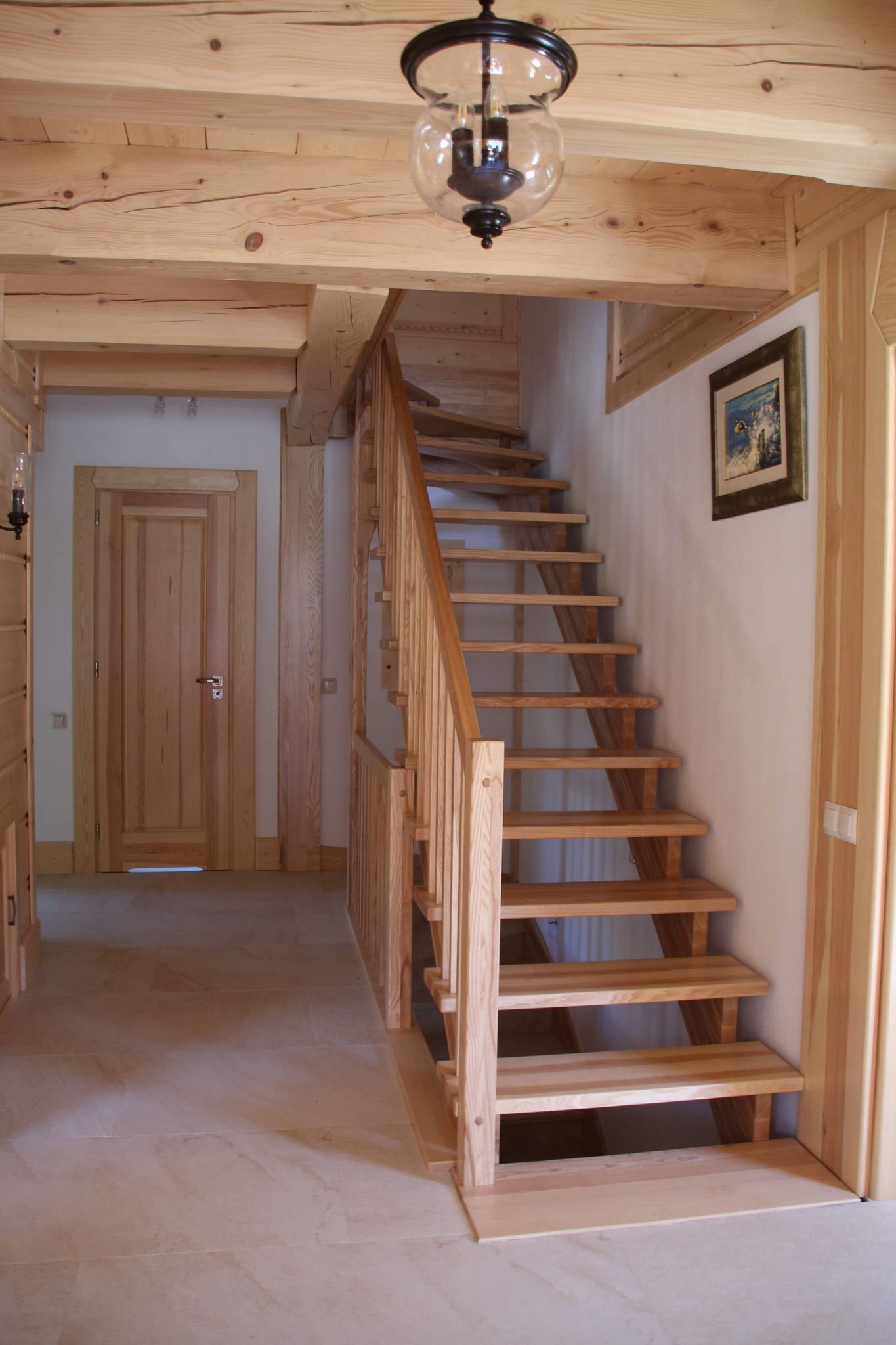 Jasne, drewniane schody z balustradą w domu o drewnianym wykończeniu, widoczne drzwi z drewnianą ramą, podłoga wyłożona jasnymi płytkami, na ścianie obraz w złotej ramie i lampa wisząca w stylu retro.