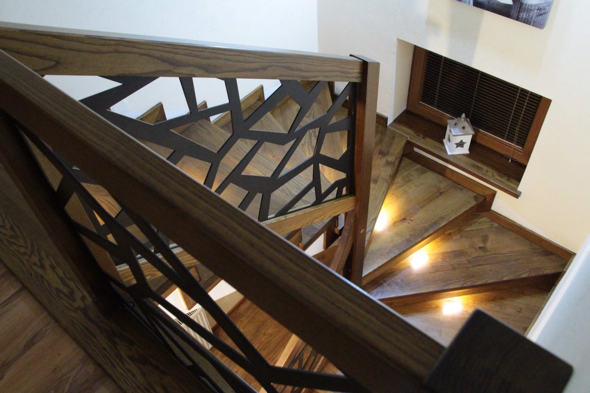 Kątowe ujęcie spiralnych schodów z dębowego drewna z metalową balustradą o geometrycznym wzorze, podświetlonych ledami, widok z góry.