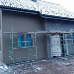 Dom jednorodzinny w trakcie prac elewacyjnych: fragment ocieplonej ściany z szarym tynkiem, rusztowania, okna zabezpieczone folią, fragment ocieplenia z płyt styropianowych, żółty przewód...