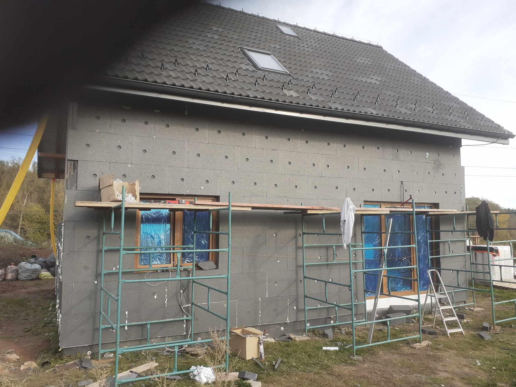 Dom jednorodzinny w trakcie ocieplania szarymi płytami styropianowymi, okna zabezpieczone folią, widoczne rusztowania i materiały budowlane.