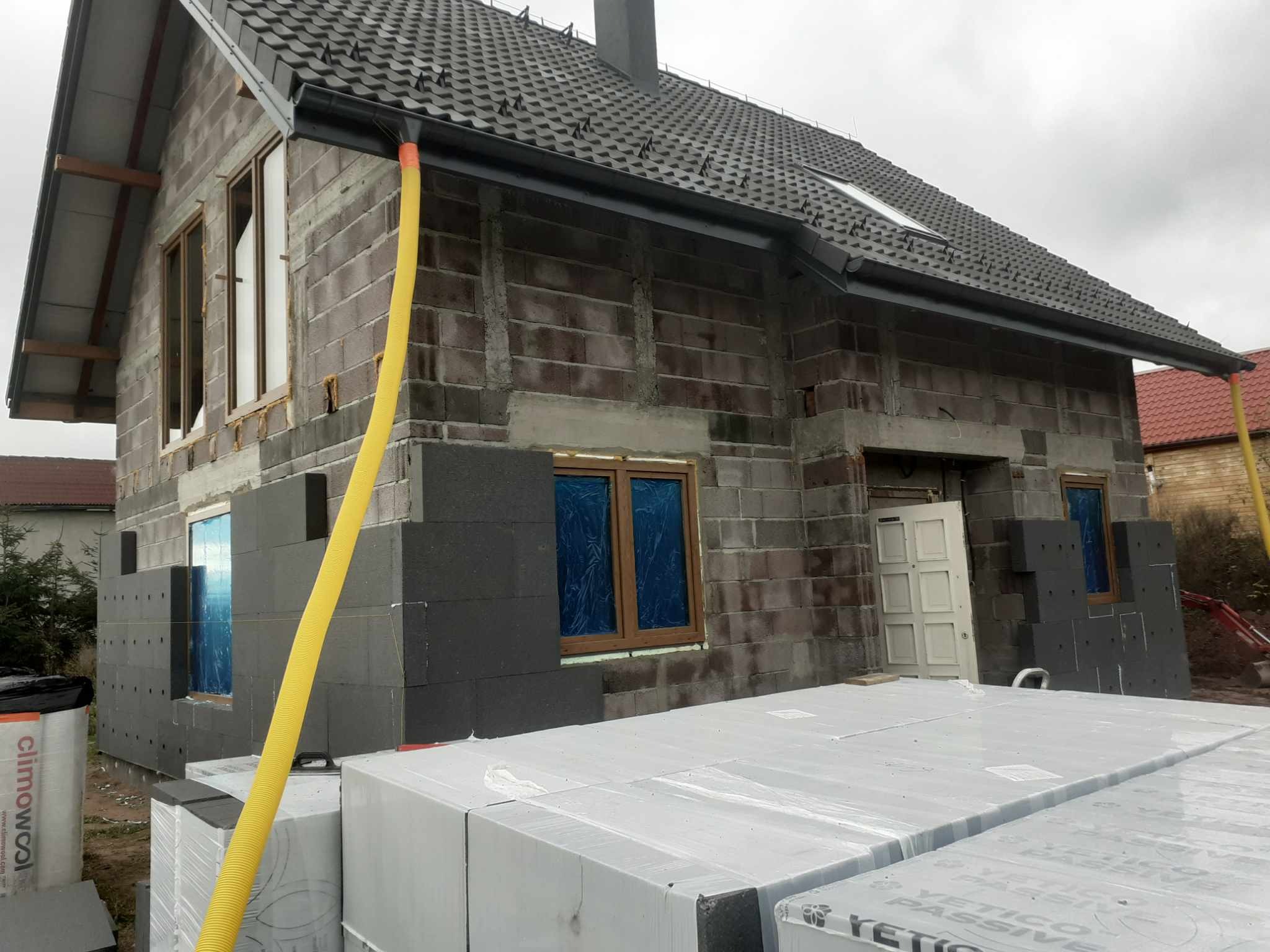 Dom w trakcie docieplania: szare bloczki, czarne płyty izolacyjne, okna zabezpieczone folią, żółty wąż wentylacyjny i palety z materiałem izolacyjnym w folii na pierwszym planie.