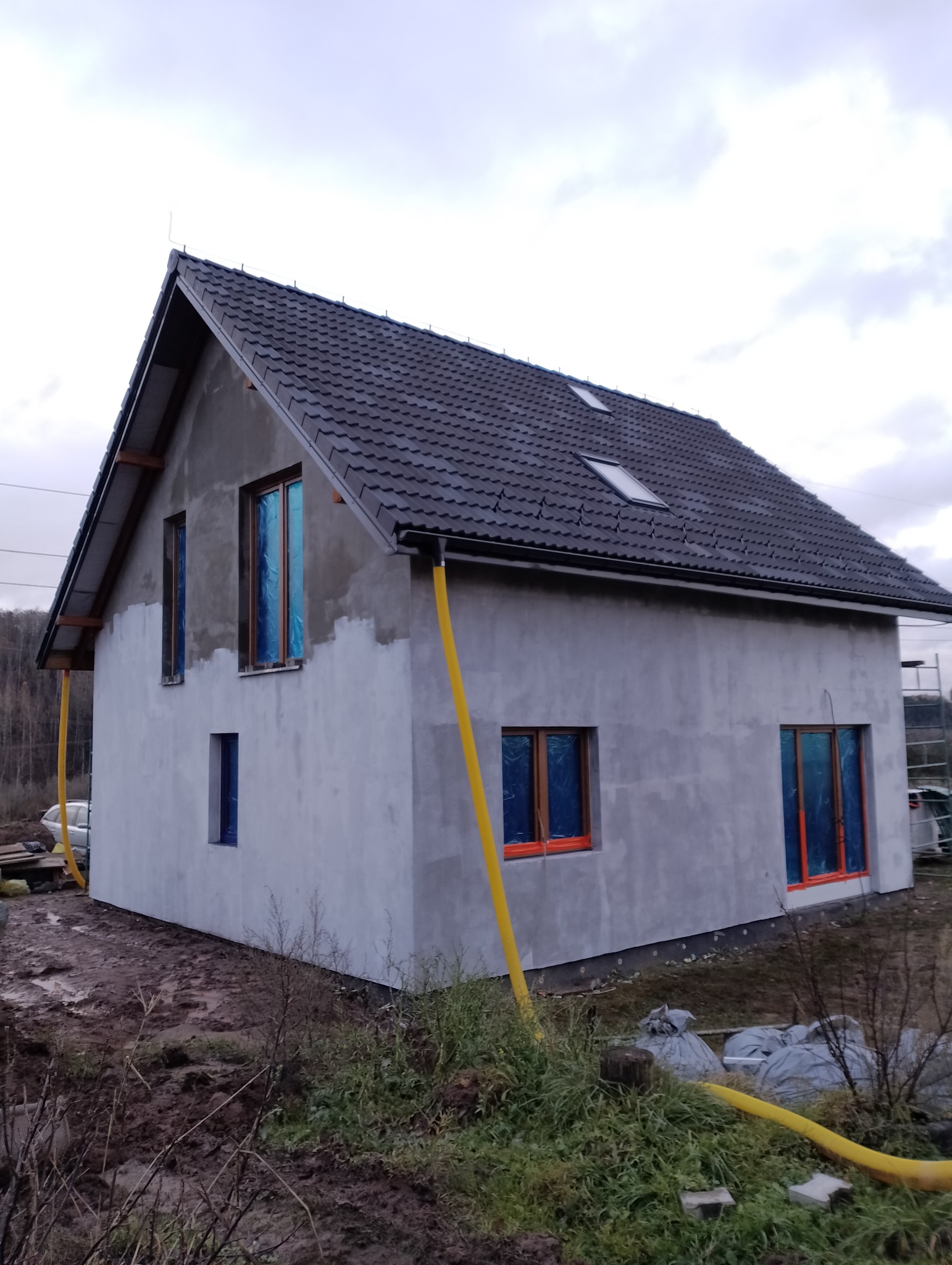 Dom jednorodzinny w trakcie tynkowania elewacji, okna zabezpieczone folią, widoczne żółte rury wentylacyjne, teren wokół błotnisty.