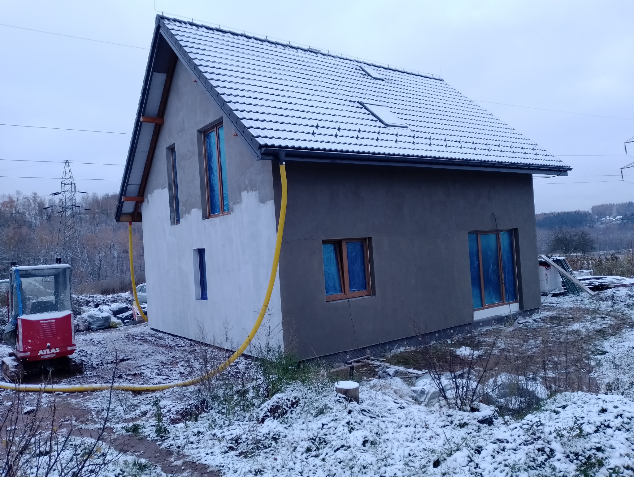 Dom w trakcie prac elewacyjnych zimą: częściowo otynkowana szara fasada, biała ściana boczna, okna zabezpieczone folią, śnieg na dachu i ziemi, żółty wąż budowlany, minikoparka Atlas w tle.