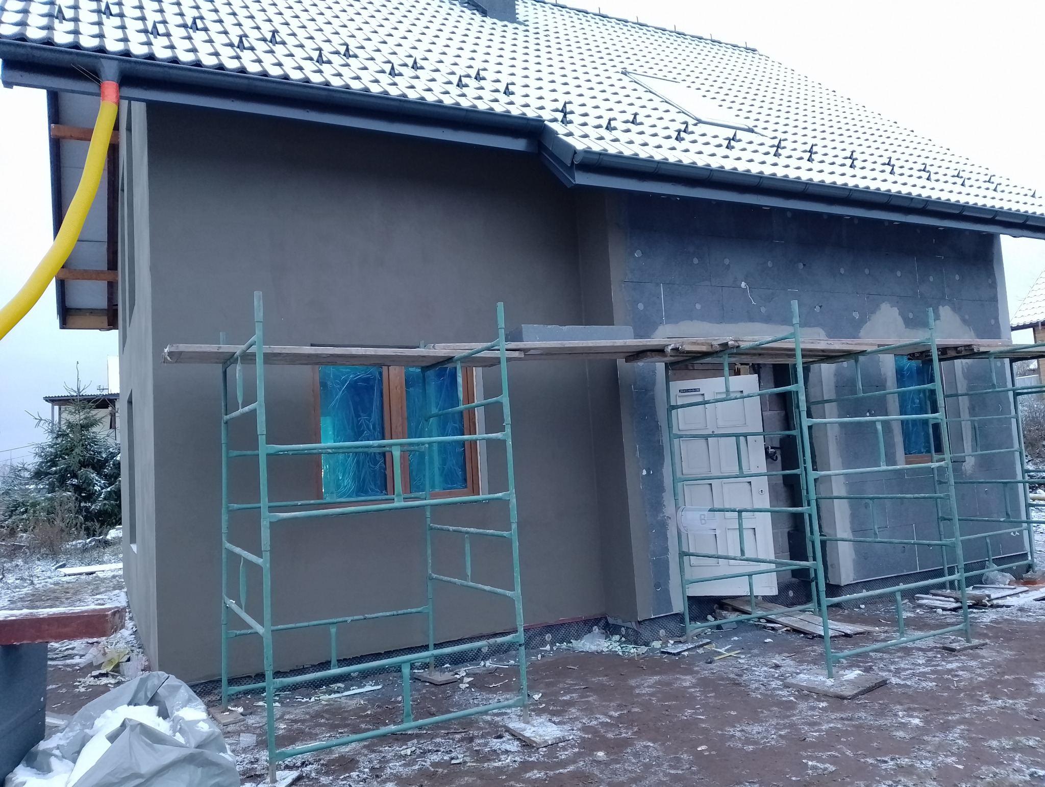 Dom jednorodzinny w trakcie prac elewacyjnych: fragment ocieplonej ściany z szarym tynkiem, rusztowania, okna zabezpieczone folią, fragment ocieplenia z płyt styropianowych, żółty przewód...