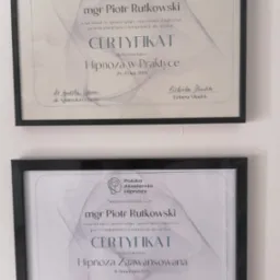 Dwa certyfikaty w czarnych ramkach wiszące na ścianie, potwierdzające ukończenie kursów z hipnozy: 'Hipnoza w Praktyce' oraz 'Hipnoza Zaawansowana' dla mgr Piotra Rutkowskiego, sygnowane przez dr...