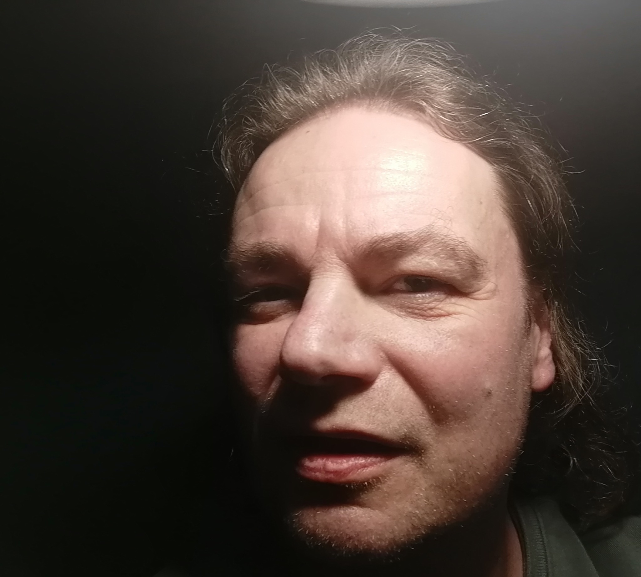 Portret mężczyzny z lekkim uśmiechem, oświetlonego z góry, z ciemnym tłem. Widoczne zmarszczki na czole i zarost.