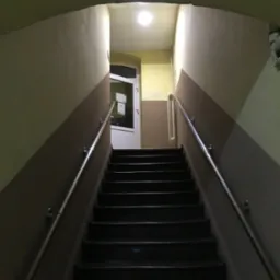 Klatka schodowa w budynku mieszkalnym z metalową poręczą, widok z dołu na g&oacute;rę schod&oacute;w, na końcu białe drzwi z szybą i oświetlenie sufitowe.