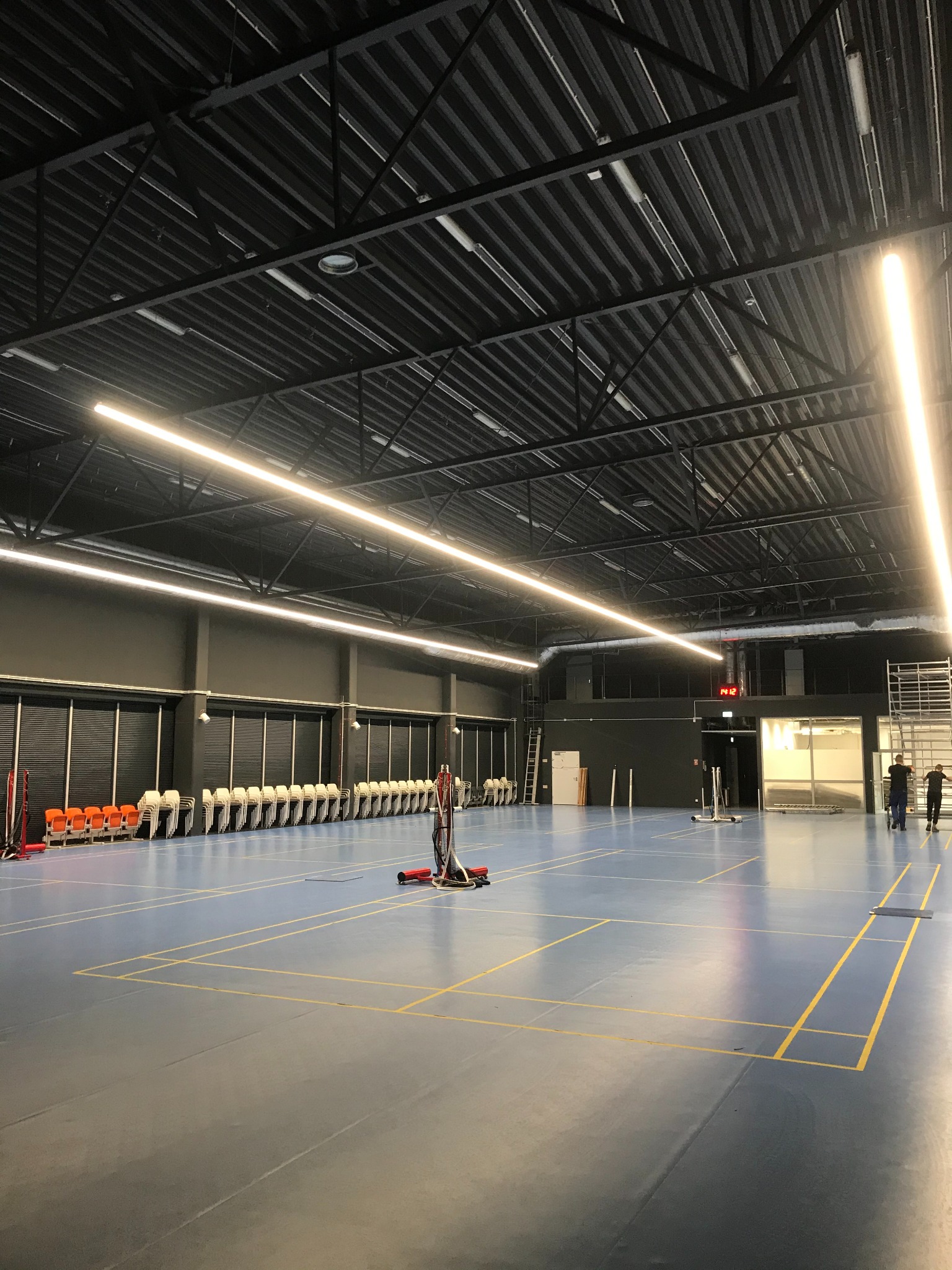 Wnętrze hali sportowej z równomiernym oświetleniem liniowym LED pod sufitem, widoczne linie boiska, rząd krzeseł pod ścianą i osoby w tle.