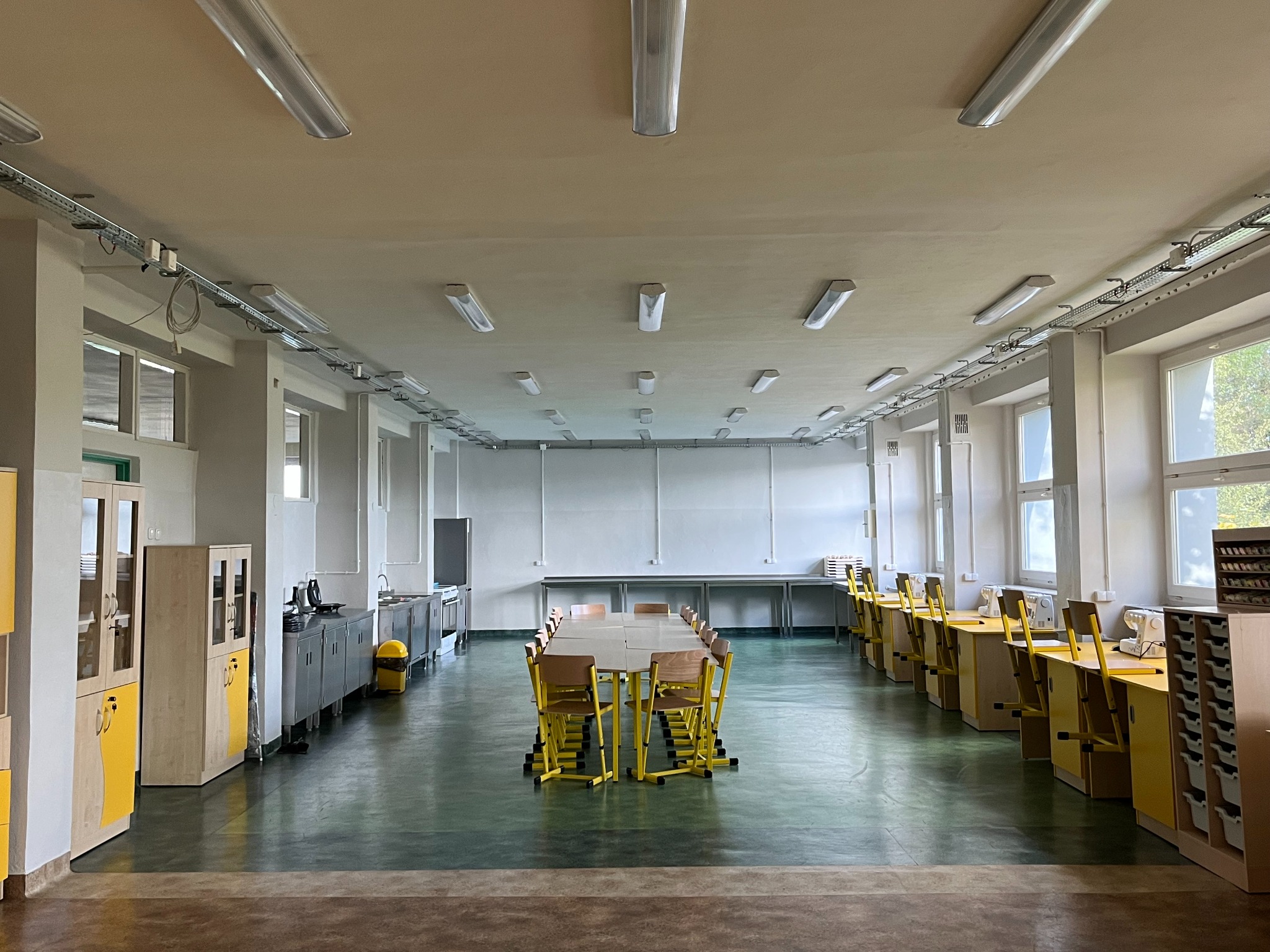 Wnętrze przestronnej sali z rzędami lamp sufitowych, widoczne koryta kablowe, stoły i krzesła w centralnej części oraz stanowiska pracy z maszynami do szycia wzdłuż ściany z oknami.