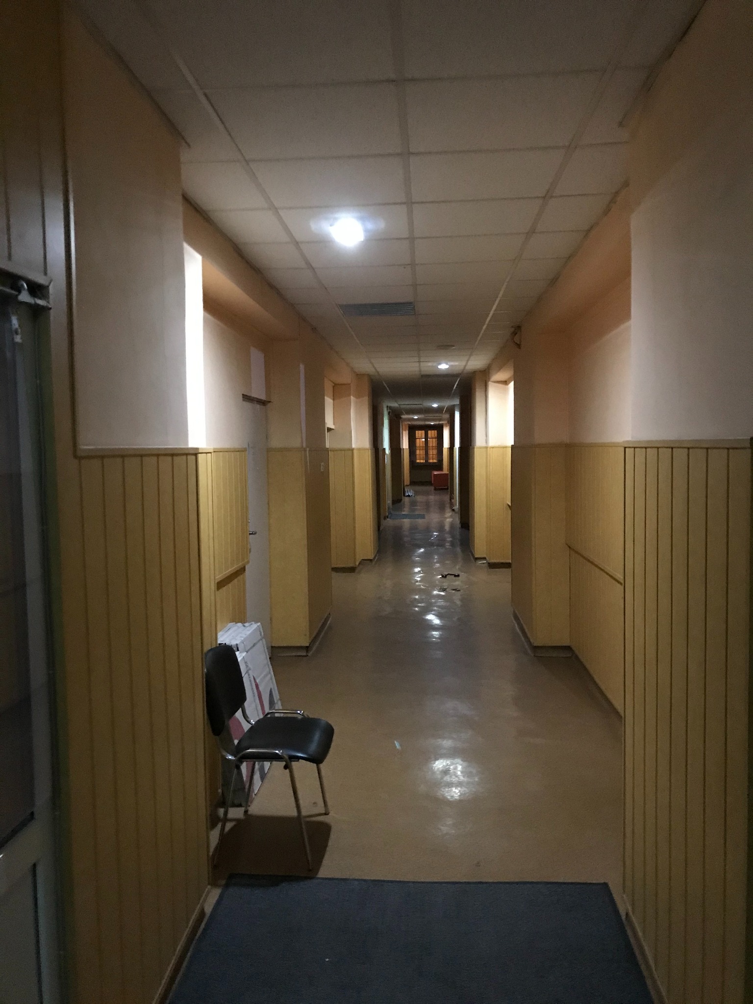 Długi, słabo oświetlony korytarz z panelami ściennymi i sufitem kasetonowym, zakończony drzwiami z widocznym światłem; krzesło biurowe i złożony karton przy ścianie po lewej stronie.