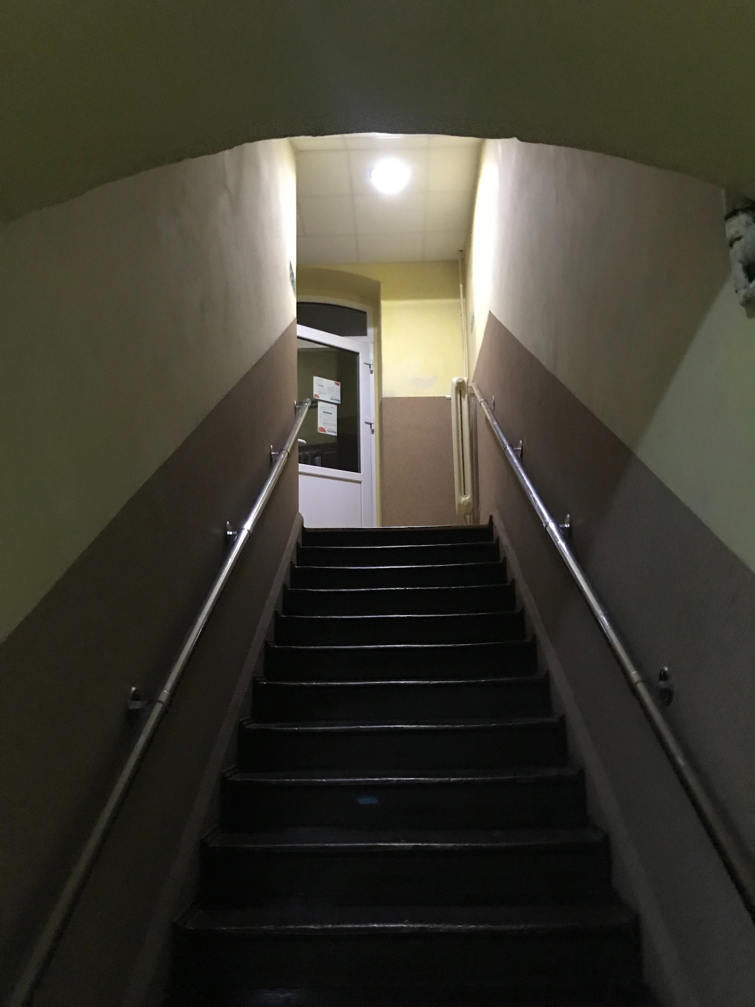 Klatka schodowa w budynku mieszkalnym z metalową poręczą, widok z dołu na górę schodów, na końcu białe drzwi z szybą i oświetlenie sufitowe.