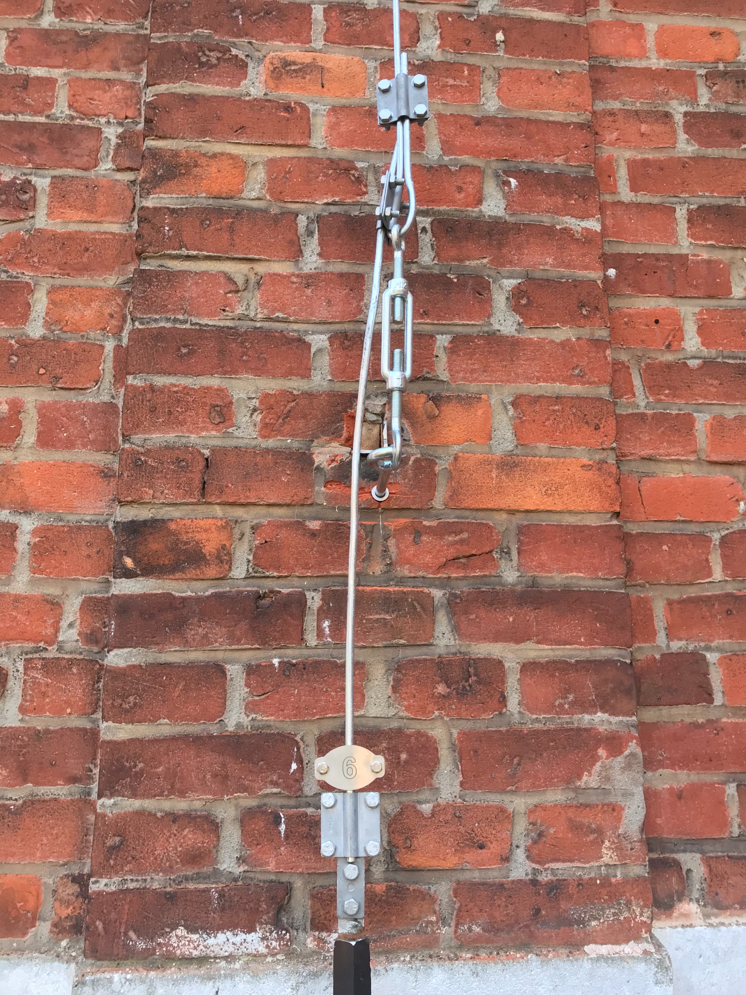 Mocowanie odgromowe na ścianie z czerwonej cegły, widoczne elementy metalowe, linka i tabliczka z numerem 6.