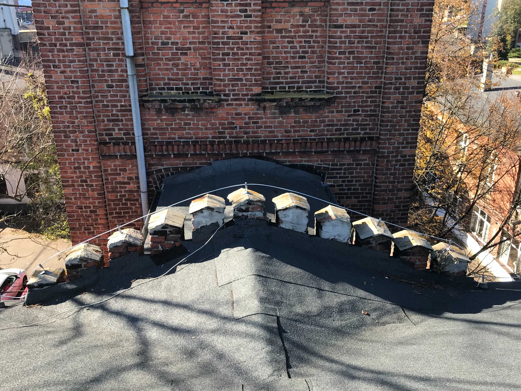 Widok z góry na dach pokryty papą z wystającymi kominami, z ceglanym budynkiem w tle i fragmentem białego samochodu po lewej stronie.