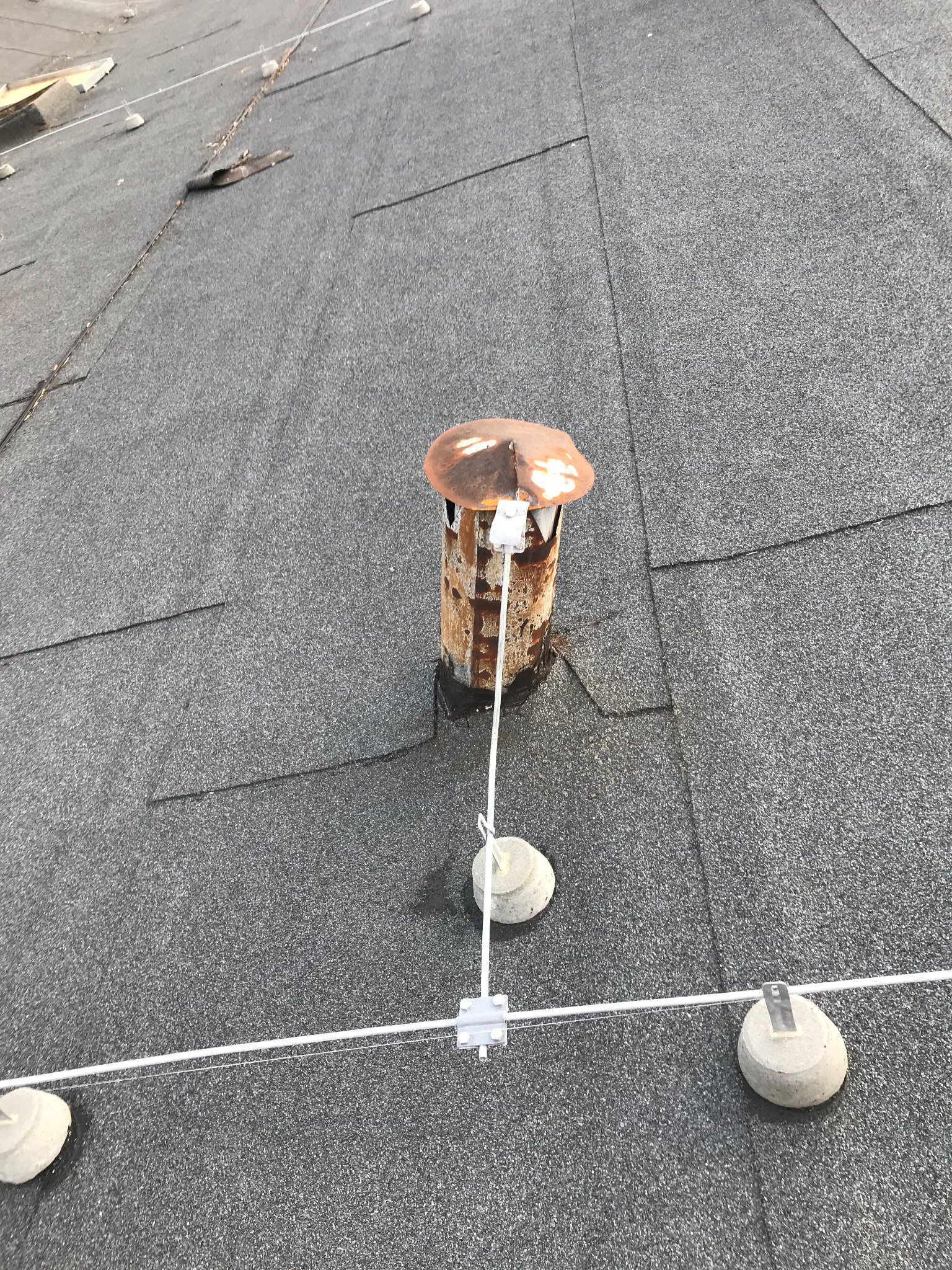 Rzut z góry na płaski dach pokryty papą, z widocznym rdzewiejącym kominkiem wentylacyjnym zabezpieczonym systemem odgromowym z betonowymi podstawami.